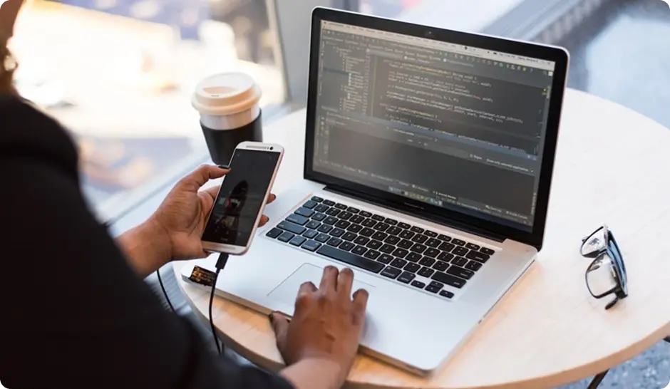 A person works on a laptop while holding a smartphone, with a coffee cup and glasses on a table nearby.